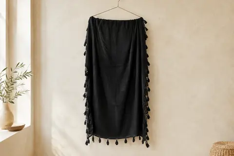 Paréo noir en coton à franges nouées, présenté seul sur un cintre dans un intérieur lumineux.